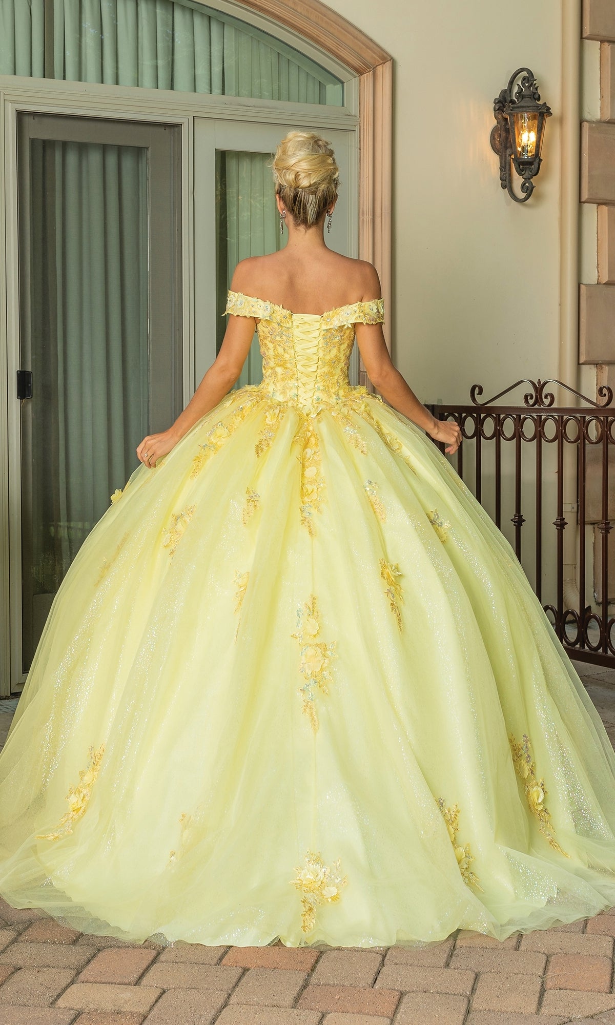 A woman stands outdoors, facing away, wearing the Quinceanera Dress 1760 By Dancing Queen—an off-the-shoulder gown with floral details and a flowing A-line ball gown silhouette.